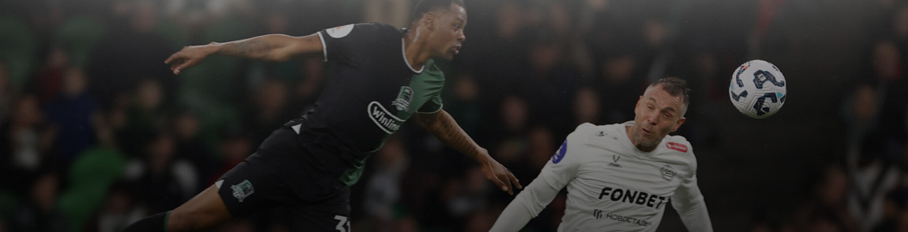 Футболист в тёмной форме прыгает за мячом против игрока в светлой форме на фоне зрителей. Оба сосредоточены на игре, мяч в воздухе