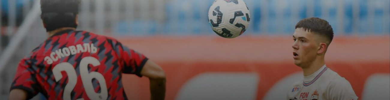 На изображении два футболиста во время игры. Один из них в красно-черной форме, другой в белой. Между ними летит футбольный мяч. На заднем плане видны трибуны с размытым изображением зрителей