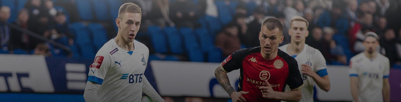 На изображении футболисты соревнуются на поле. Игрок в белой форме противостоит игроку в чёрно-красной форме. На заднем плане можно увидеть других спортсменов и зрителей на трибунах