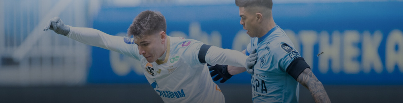 Футболист в белой форме борется за мяч с игроком в голубой форме на поле. На заднем плане видны размытые трибуны и рекламный баннер