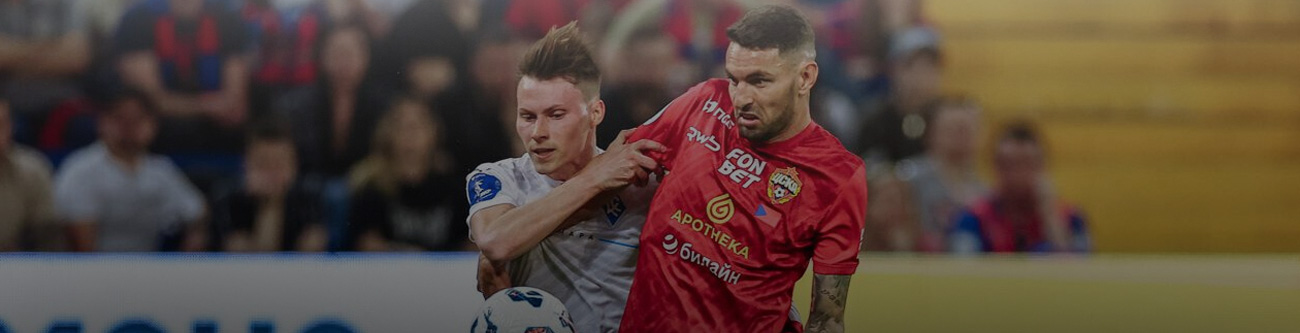 На изображении два футболиста: один в серой форме, другой в красной. Они борются за мяч на фоне трибун с болельщиками