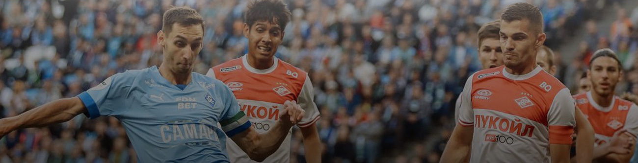 Футбольный матч: игрок в голубой форме борется за мяч с двумя соперниками в красно-белой форме. На заднем плане размытая толпа зрителей на трибунах