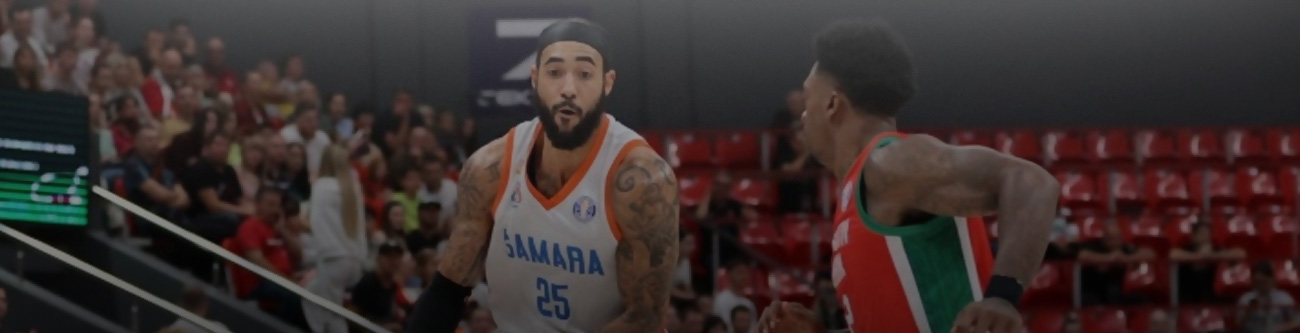 Баскетбольный матч: игрок в белой форме с номером 25 смотрит в сторону партнёра по команде, в то время как его преследует соперник в зелёно-красной форме. На заднем плане видны зрители на трибунах