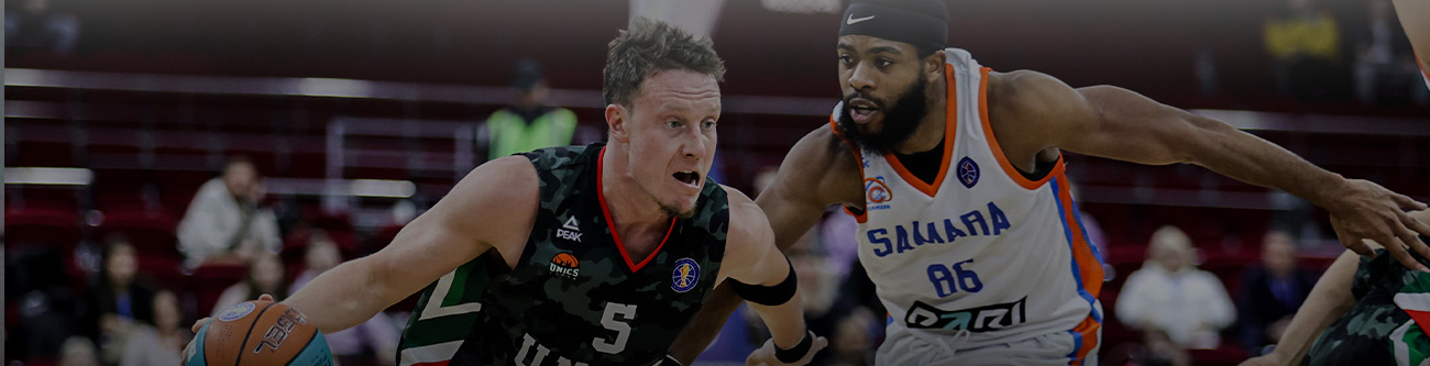 На изображении показан динамичный момент баскетбольного матча. Игрок в тёмной форме ведёт мяч, а соперник в белой форме пытается его остановить. Позади видны зрители и часть баскетбольной площадки